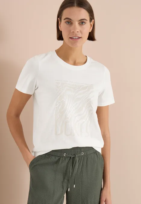 Eine Frau trägt ein weißes T-Shirt mit dezentem Print und eine olivgrüne, weite Hose.