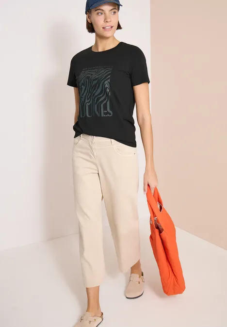 Frau trägt schwarzes T-Shirt, helle weite Hose und hält eine orangene Tasche.