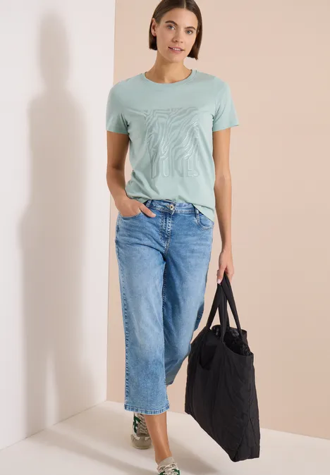 Frau steht in hellem Raum, trägt hellgrünes T-Shirt, blaue Jeans mit Umschlag und hält eine große schwarze Tasche.