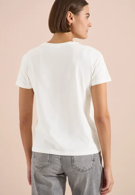 T-shirt à col rond et imprimé sur le devant vanilla white Frau steht vor hellem Hintergrund, trägt weißes T-Shirt und graue Jeans, Rückenansicht.