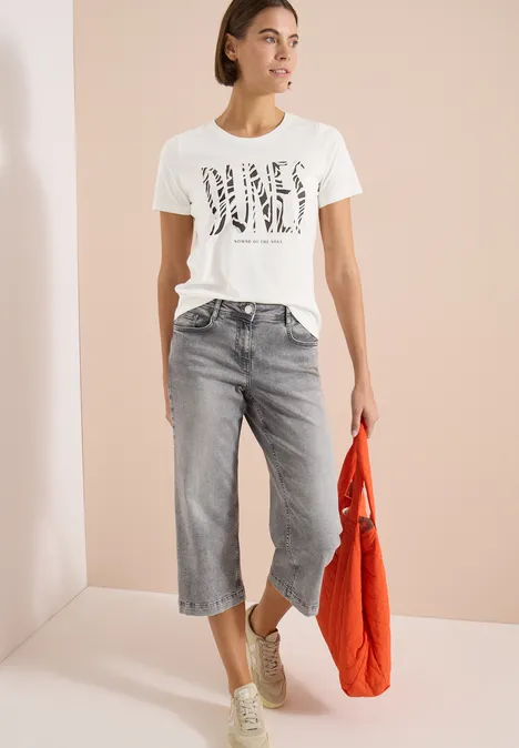 T-shirt à col rond et imprimé sur le devant vanilla white Frau im weißen Print-T-Shirt, grauen Culotte-Jeans, Sneakern und orangefarbener Tasche.