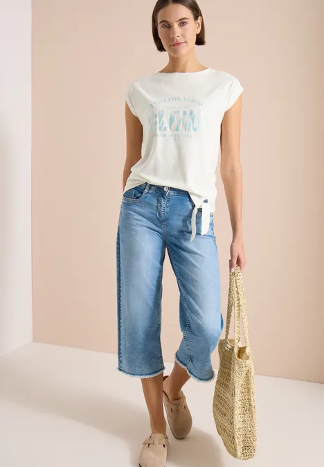 Vrouw met wit T-shirt, wijde culotte-jeans, strooien tas en espadrilles tegen een lichte achtergrond.