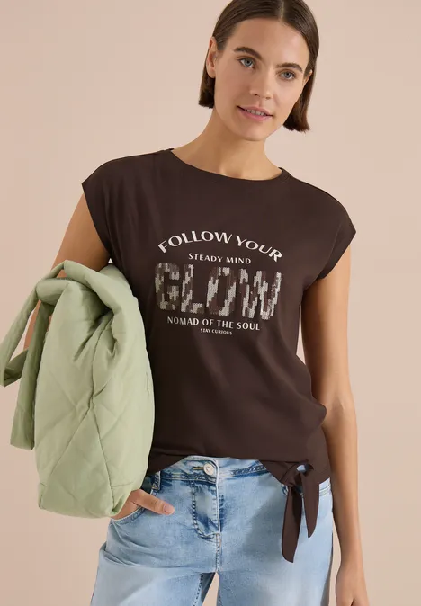 Frau im schwarzen, ärmellosen Top mit Aufdruck und seitlicher Schleife, in Jeans, grüne Jacke über dem Arm.