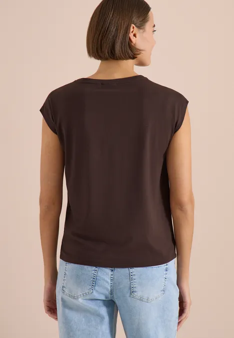Frau von hinten in dunklem T-Shirt mit Schulterpolstern und blauer Jeans vor beigem Hintergrund.