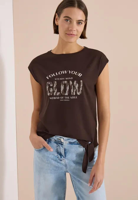 Frau in braunem, seitlich geknotetem T-Shirt mit Aufdruck und hellen Jeans, locker stehend mit einer Hand in der Tasche.