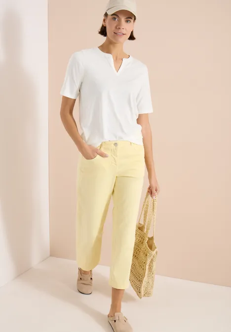 Frau trägt weißes V-Shirt, helle gelbe Hose und beige Sneaker, hält geflochtene Strohtasche vor hellem Hintergrund.