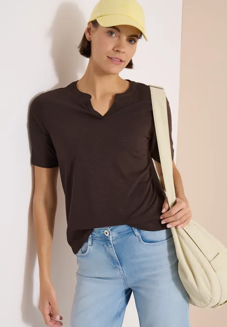 Frau in dunklem T-Shirt, hellblauen Jeans, hellbeigem Schulterbeutel und Cap vor zweifarbiger Wand