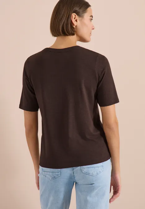 Frau von hinten in braunem T-Shirt mit Halbarm und hellblauer Jeans vor beigem Hintergrund.