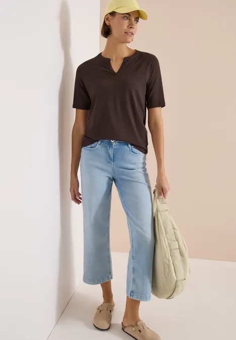 Frau in schwarzem V-Shirt, hellen Jeans, beigen Slip-ons und großer Tasche vor neutralem Hintergrund.
