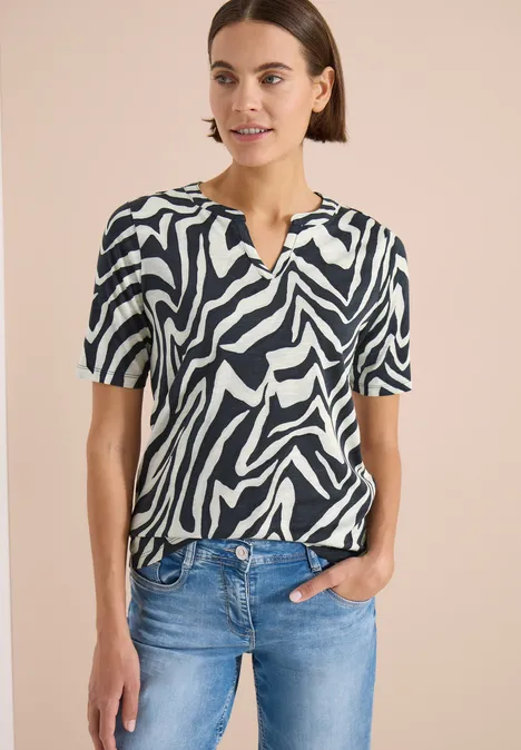 Vrouw in zwart-wit zebraprint T-shirt met korte mouwen en blauwe spijkerbroek, hand in zak.