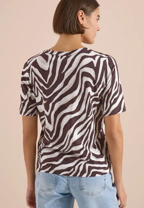 Iemand van achteren in een zwart-wit zebraprint t-shirt en een lichtblauwe spijkerbroek voor een beige achtergrond.