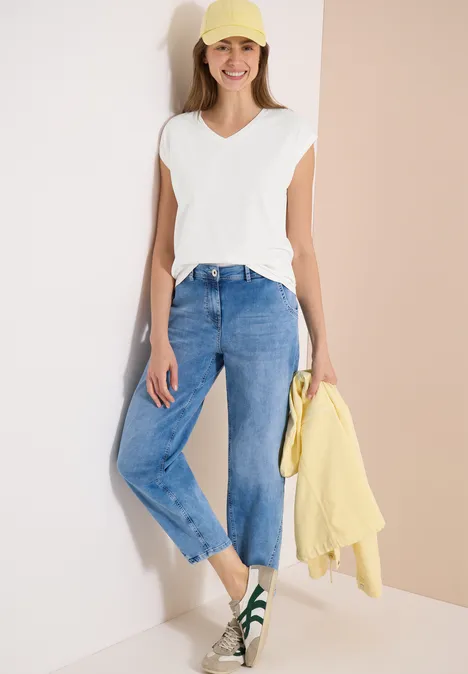 Frau mit weißem T-Shirt, lockeren blauen Jeans und grünen Sneakern, hält eine gelbe Jacke.