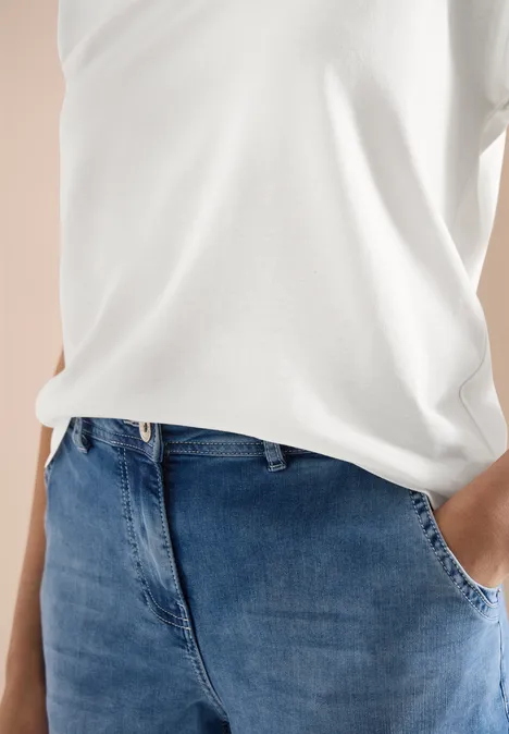 Eine Person trägt ein weißes T-Shirt und blaue Jeans, eine Hand steckt in der Hosentasche.