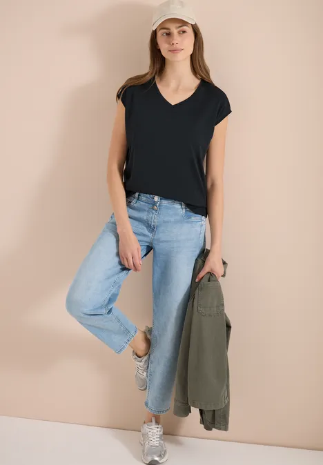 Frau lehnt an hellem Hintergrund, trägt schwarzes T-Shirt, blaue Jeans, weiße Sneakers und hält eine grüne Jacke.