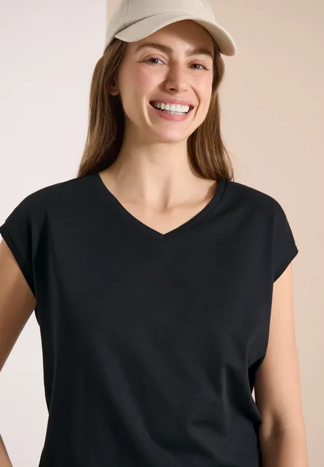 Frau mit schwarzem V-Neck-Shirt und hellbeiger Baseballkappe vor neutralem Hintergrund.