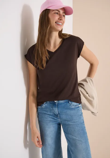Vrouw in bruin T-shirt, blauwe jeans en roze pet, tegen een lichte muur geleund, jas over de arm.