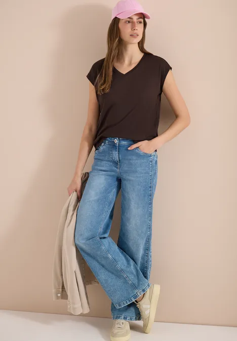 Vrouw in zwart T-shirt, blauwe spijkerbroek en sneakers, met een beige jas in de hand.