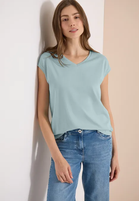 Eine Frau lehnt an einer Wand und trägt ein hellblaues T-Shirt sowie Jeans.
