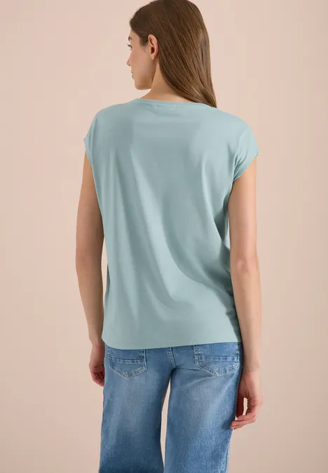 Frau von hinten in hellblauem lockerem T-Shirt und blauen Jeans vor neutralem Hintergrund.