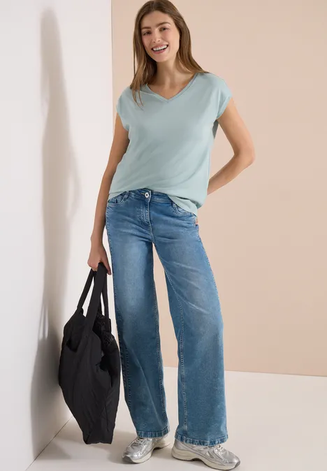 Eine Frau lehnt an einer Wand, trägt hellblaues T-Shirt, weite Jeans, weiße Sneaker und hält eine schwarze Tasche.