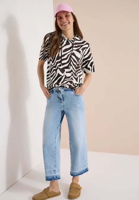 Frau trägt Zebra-Bluse, helle weite Jeans und braune Sandalen, steht vor neutralem Hintergrund.