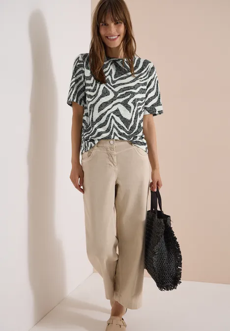 Vrouw in zebraprint top, losse beige broek en zwarte tas voor een lichte muur.