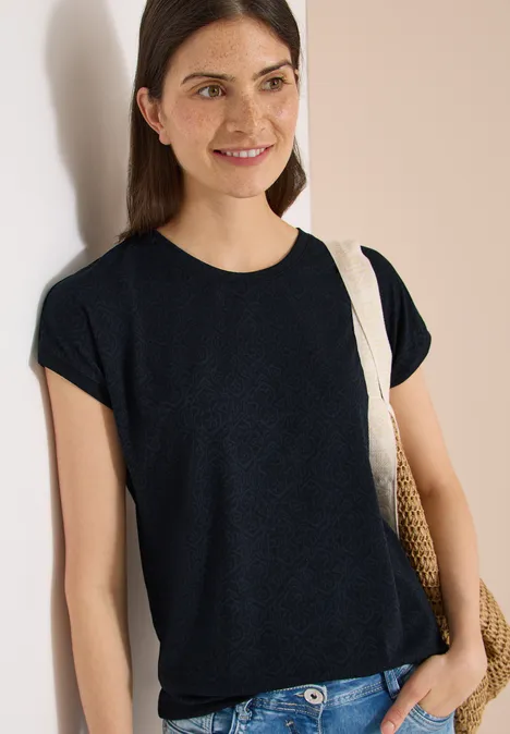 Person lehnt an Wand, trägt schwarzes T-Shirt und Strohschultertasche.