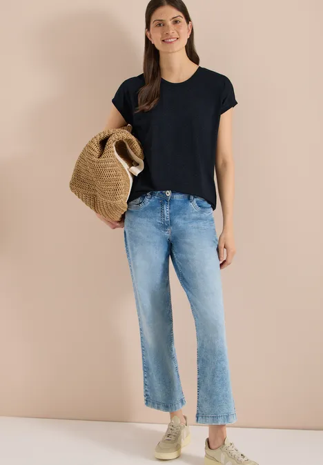 Frau vor beiger Wand in schwarzem T-Shirt, hellen Jeans, Sneakers und mit geflochtenem Strohkörbchen.