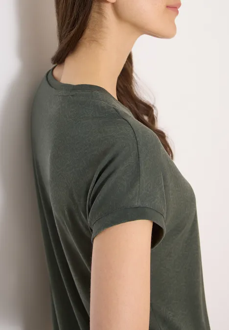 Junge Frau seitlich, lehnt an weißer Wand, trägt dunkelgrünes kurzärmeliges T-Shirt, Blick nach vorn