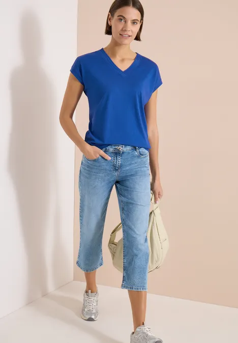 Frau trägt blaues V-Shirt, Capri-Jeans und weiße Sneakers, hält eine helle Tasche.