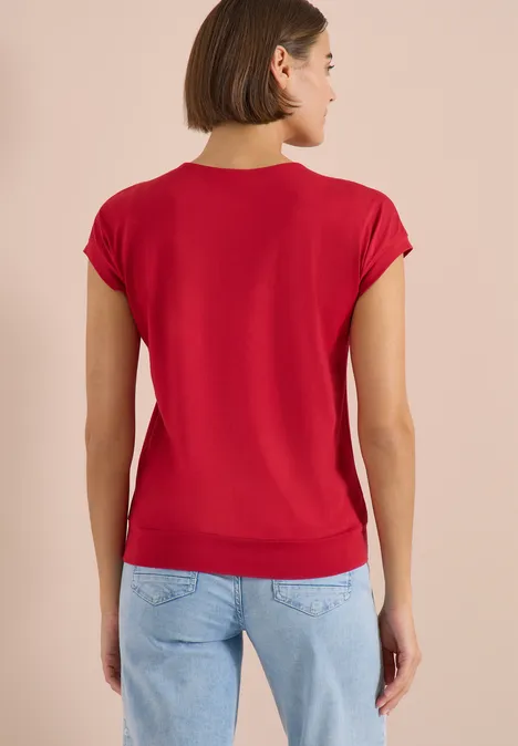 T-shirt à manches courtes et col en V en couleur unie tamarillo red Femme vue de dos portant un t-shirt rouge et un jean bleu clair sur fond beige