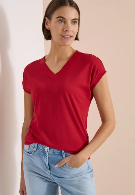 T-shirt à manches courtes et col en V en couleur unie tamarillo red Femme en T-shirt rouge à col V et jean bleu, debout les mains dans la poche devant un fond sobre.