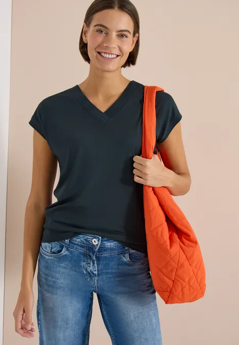 Frau trägt dunkelblaues V-Neck-Shirt, Jeans und hält eine orange gesteppte Tasche.