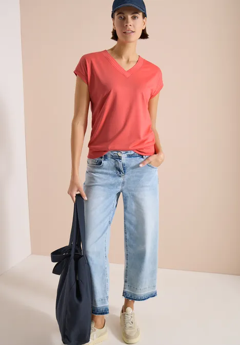 Persoon voor een beige achtergrond, draagt een rood t-shirt, lichte jeans, sneakers en houdt een tas vast.