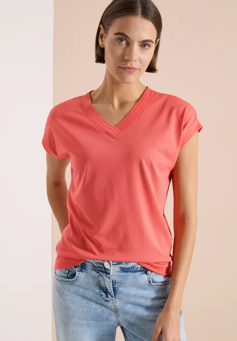 Vrouw draagt koraalkleurig V-hals T-shirt en spijkerbroek voor tweekleurige achtergrond.