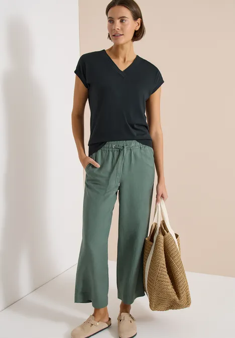 Eine Frau trägt ein schwarzes T-Shirt, locker geschnittene grüne Hose und hält eine große geflochtene Tasche.