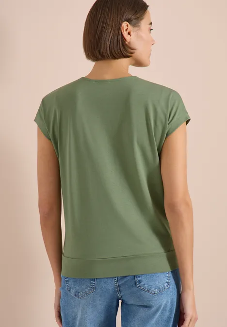Frau von hinten in olivgrünem T-Shirt und blauer Jeans vor neutralem Hintergrund.