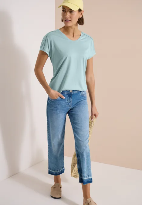 T-Shirt mit V-Neck und Spitzendetail crystal mint Frau trägt helltürkisfarbenes V-Shirt, lockere blaue Jeans und beige Slipper vor neutralem Hintergrund.