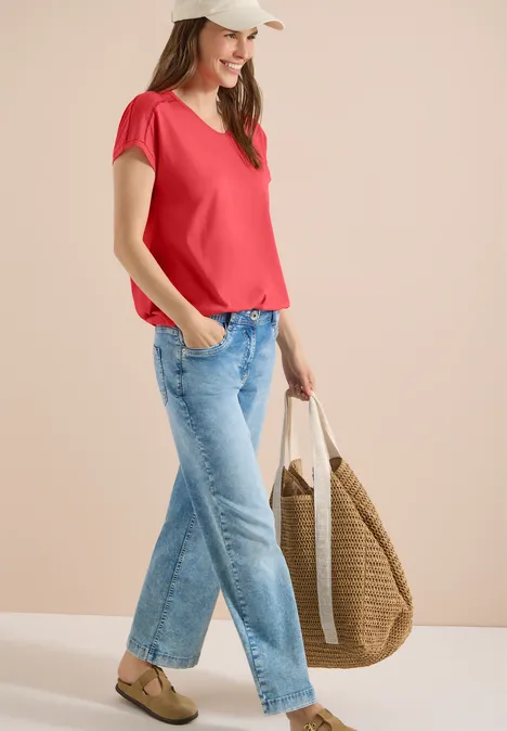 T-Shirt mit V-Neck und Spitzendetail coral Frau in rotem T-Shirt, blauen Jeans und großer geflochtener Tasche vor neutralem Hintergrund.