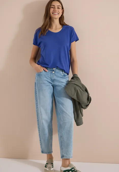 Shirt met V-hals en kanten detail deep river blue Vrouw in blauw t-shirt en lichtblauwe jeans, houdt een groene jas vast voor een beige achtergrond.