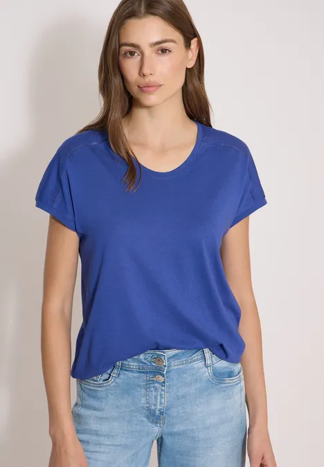 Shirt met V-hals en kanten detail deep river blue Vrouw draagt een blauw T-shirt en spijkerbroek, staand voor een neutrale achtergrond.