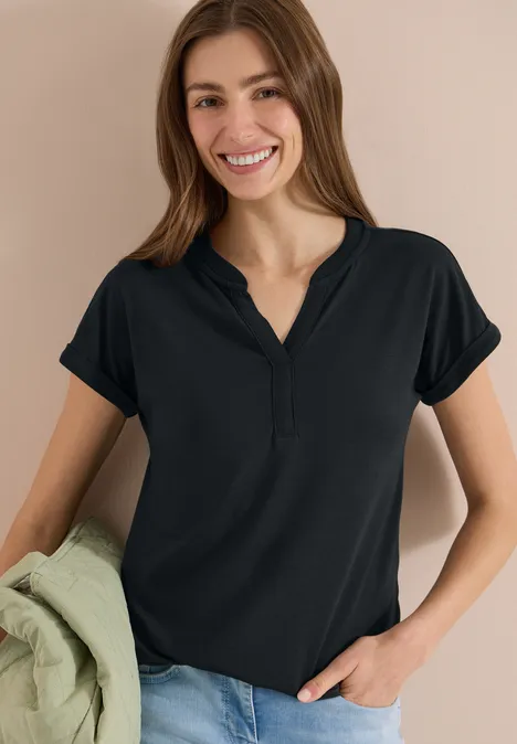 Frau in schwarzem Kurzarmshirt und Jeans, hält eine grüne Jacke lässig in der Hand.