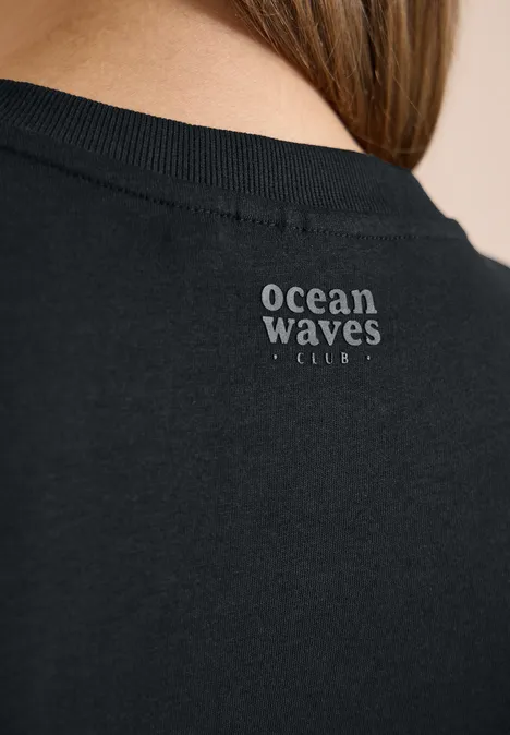 Schwarzes T-Shirt mit weißem Schriftzug 'ocean waves club' am Nackenbereich.