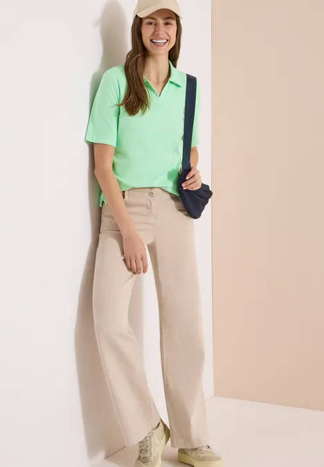 Frau lehnt an heller Wand, trägt mintgrünes Polo, beige weite Hose, weiße Sneaker und schwarze Umhängetasche.