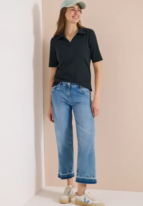 Frau in schwarzem Poloshirt, hellblauen Cropped-Jeans, Kappe und Sneakers vor beigem Hintergrund.