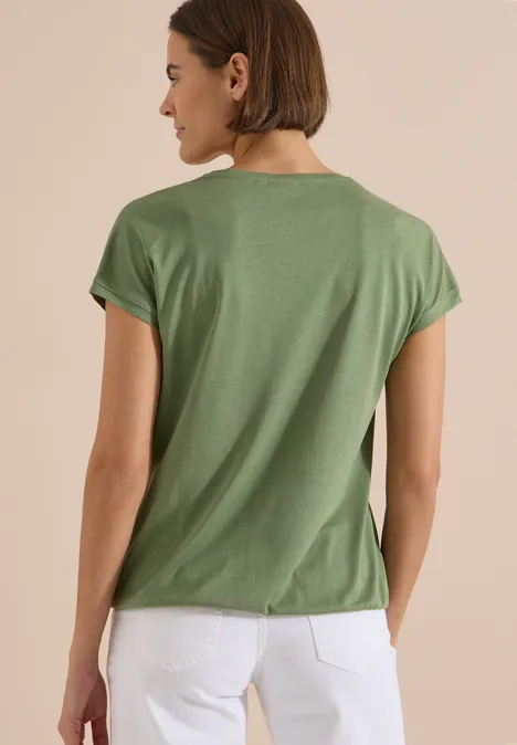 T-Shirt mit V-Neck und Spitzendetail deep matcha green Eine Frau von hinten in grünem T-Shirt und weißer Hose vor schlichtem Hintergrund.