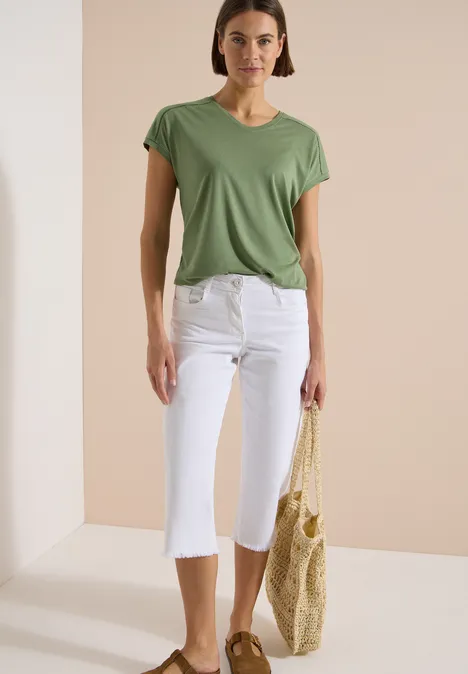 T-Shirt mit V-Neck und Spitzendetail deep matcha green Frau trägt grünes T-Shirt, weiße Capri-Hosen und hält eine geflochtene Strohtragetasche.