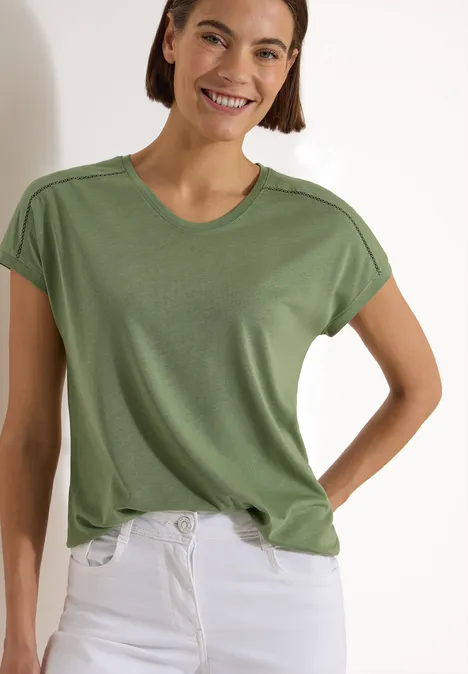 T-Shirt mit V-Neck und Spitzendetail deep matcha green Eine Person trägt ein schlichtes grünes T-Shirt und weiße Jeans, Oberkörper sichtbar.