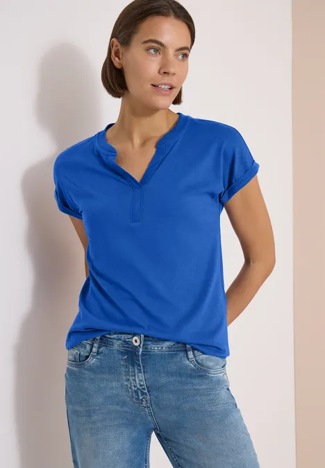 Kurzarm Shirt im Tunika-Look surf blue Person mit kurzen Haaren trägt blaues Polo-Shirt und Jeans, steht vor hellem Hintergrund, Gesicht unscharf.