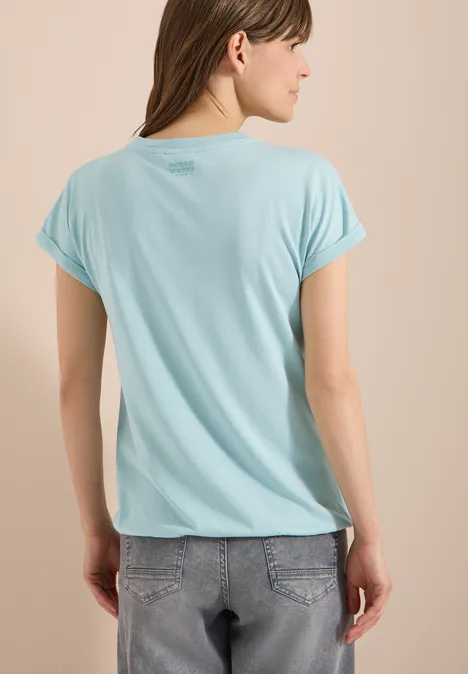 Kurzarm Shirt im Tunika-Look crystal mint Frau von hinten in hellblauem T-Shirt und grauen Jeans vor beigem Hintergrund, Gesicht unscharf.